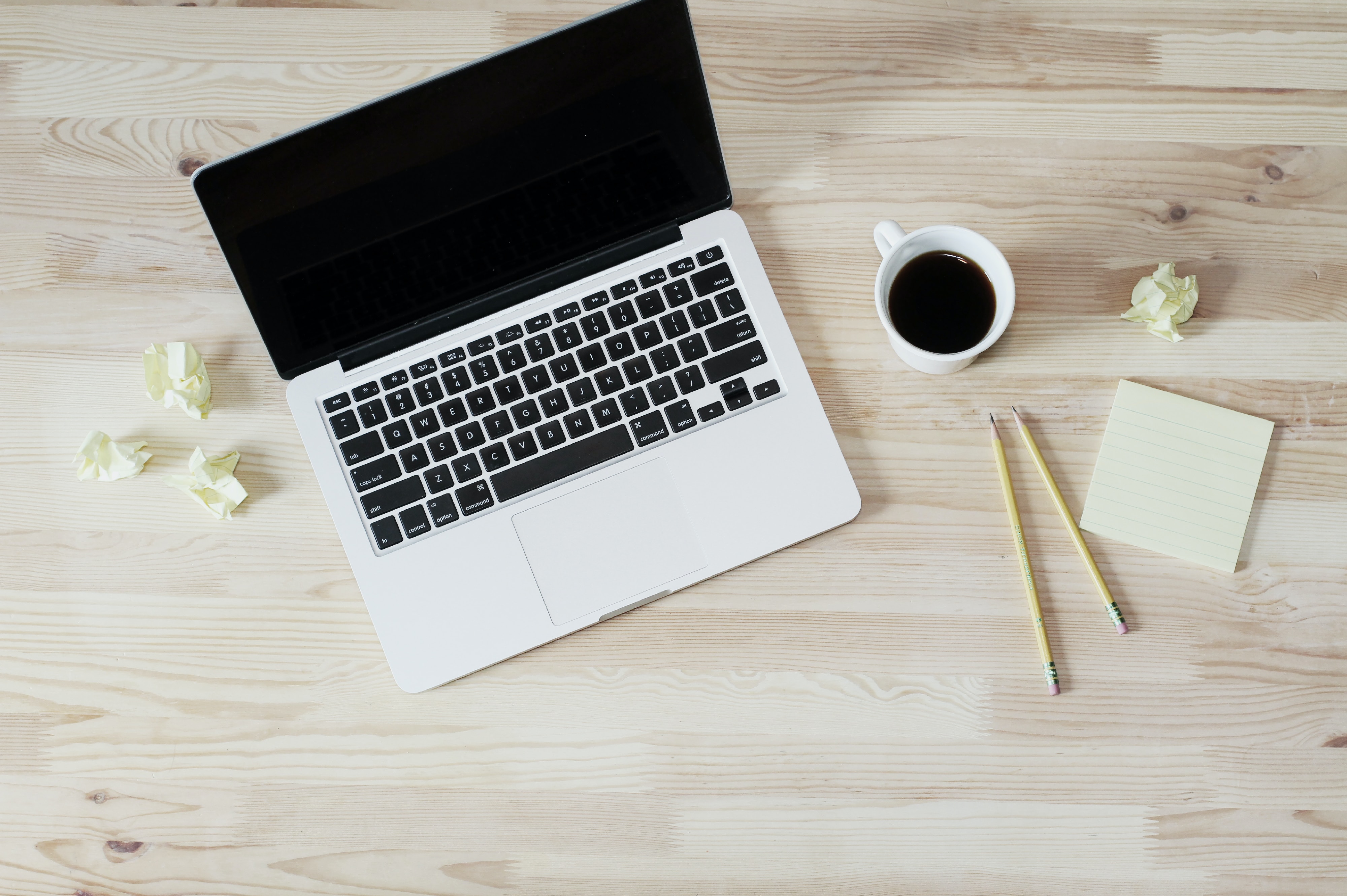 Open laptop on table next to cup of coffee and a notepad and pencil